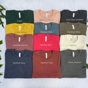 K&ouml;nnte beinhalten: Eine Sammlung von zw&ouml;lf T-Shirts mit Rundhalsausschnitt, die in einem Raster angeordnet sind und die Farben Forest, Heather Peach, Dark Grey Heather, Maize Yellow, Heather Slate, Heather Olive, Heather Mauve, Heather Red, Natural, Heather Navy, Heather Clay und Deep Heather tragen.