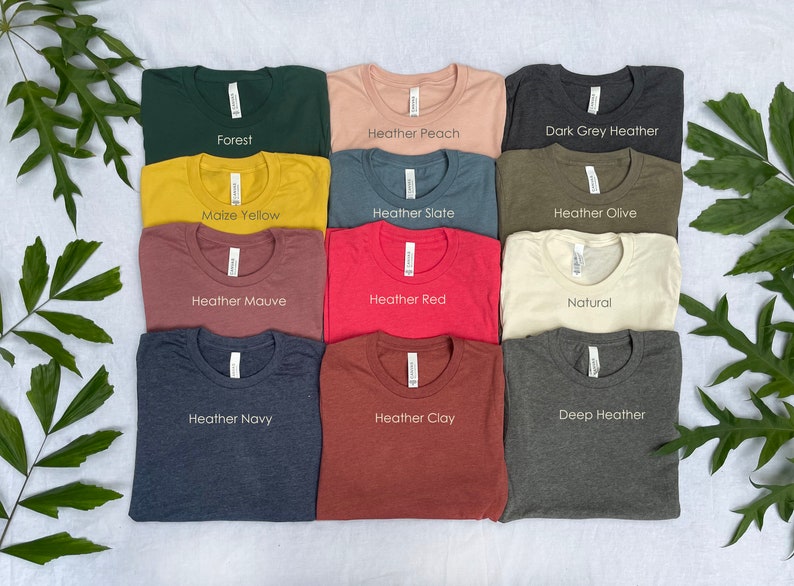 K&ouml;nnte beinhalten: Eine Sammlung von zw&ouml;lf T-Shirts mit Rundhalsausschnitt, die in einem Raster angeordnet sind und die Farben Forest, Heather Peach, Dark Grey Heather, Maize Yellow, Heather Slate, Heather Olive, Heather Mauve, Heather Red, Natural, Heather Navy, Heather Clay und Deep Heather tragen.