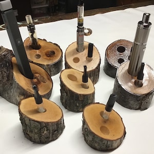 May include: A set of eight wooden log holders for vape pens and e-cigarettes. The holders are made from natural wood and have a rustic look. Each holder has a hole in the center to hold a vape pen or e-cigarette.