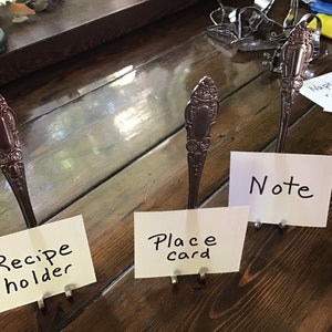 May include: Three silver spoons bent into holders for cards or notes. The spoons are on a wooden surface with handwritten notes that say "Recipe holder", "Place card", and "Note".