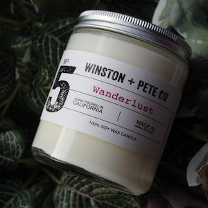 May include: A soy wax candle in a glass jar with a silver lid. The label reads "WINSTON + PETE CO Wanderlust" and "Hand Poured in California." The candle is surrounded by green foliage.