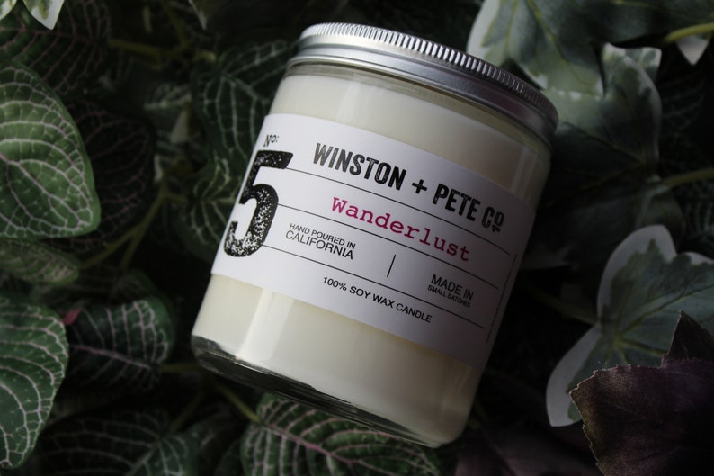 May include: A soy wax candle in a glass jar with a silver lid. The label reads "WINSTON + PETE CO Wanderlust" and "Hand Poured in California." The candle is surrounded by green foliage.
