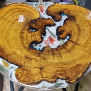 May include: A wooden table top with a natural wood grain pattern and a clear epoxy resin coating. The resin contains small, colorful fish and other decorative elements.