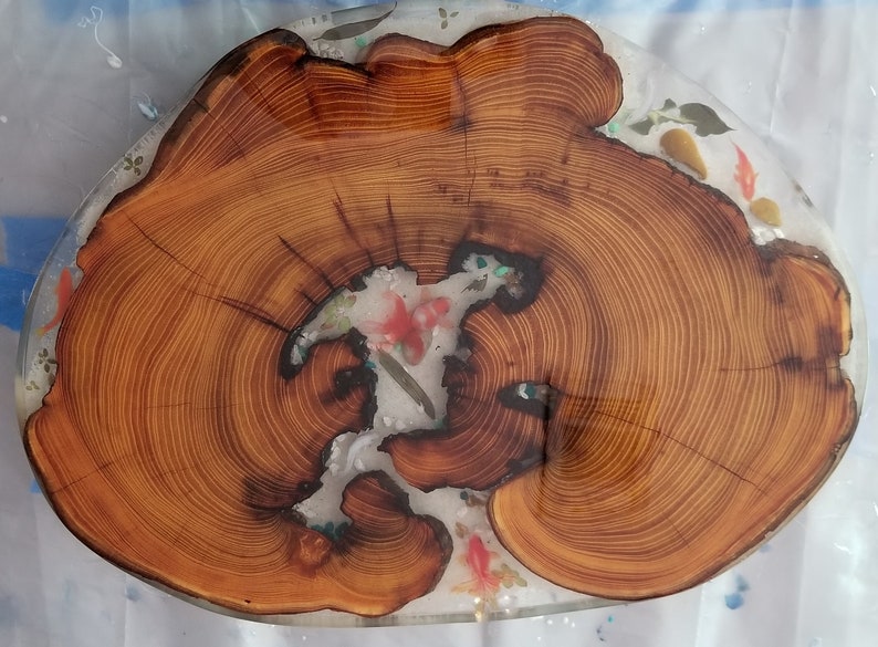 May include: A wooden slice with a natural wood grain pattern, filled with clear resin and embedded with colorful flowers and fish. The resin is clear and glossy, and the flowers and fish are visible through the resin.