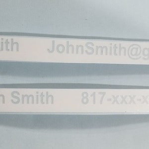 以下が含まれることがあります： 黒い文字で書かれた白いラベルが2つあります。 上のラベルには「John Smith」と「JohnSmith@gmail.com」と書かれています。 下のラベルには「John Smith」と「817-XXX-XXXX」と書かれています。