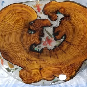 May include: A round wooden slice with a natural wood grain pattern, encased in clear resin. The resin contains embedded colorful fish and other decorative elements.