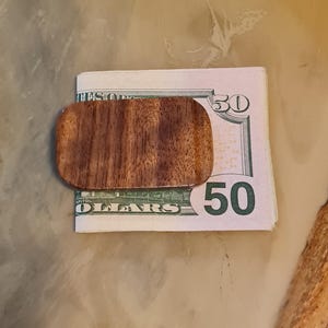 May include: A wooden money clip with a natural wood grain pattern. The clip is holding a $50 bill.