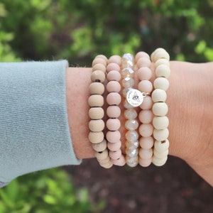 Könnte beinhalten: Ein Set aus fünf Armbändern, darunter vier Armbänder aus Holzperlen und ein Kristallarmband. Die Holz-Armbänder sind hellbeige und das Kristallarmband ist klar mit einem leichten Schimmer. Die Armbänder sind an einem Handgelenk übereinander getragen und haben einen kleinen goldenen Anhänger mit einem Bienenmotiv.