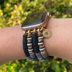 May include: A gold Apple Watch with a black face and a black, white, and gray beaded bracelet. The bracelet has a gold charm with a bee and the text "JORDYN'S SWEETS".