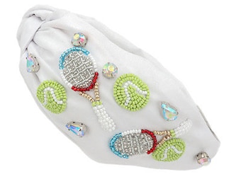 May include: White headband featuring beaded tennis rackets and tennis balls. The headband is made from a soft, white fabric and has a knotted design at the back.
