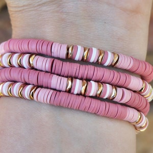 May include: A set of four pink and white beaded bracelets with gold accents. The bracelets are stacked on a person's wrist.