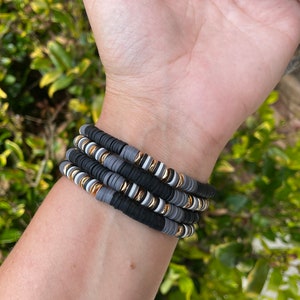 May include: A stack of four bracelets made of black, gray, and white beads with gold accents. The bracelets are stacked on a person's wrist.