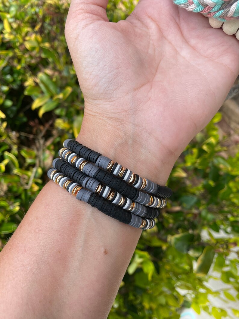 May include: A stack of four bracelets made of black, gray, and white beads with gold accents. The bracelets are stacked on a person's wrist.