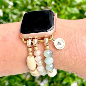 May include: A gold Apple Watch with a beaded band. The band is made of white, beige, and light blue beads. A gold charm with a bee and the text "JORDYN JAMES" hangs from the band.