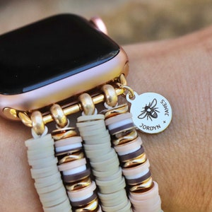 May include: A gold Apple Watch with a beige and brown beaded band. The band has a charm with a bee and the text "JAMES x JORDYN".