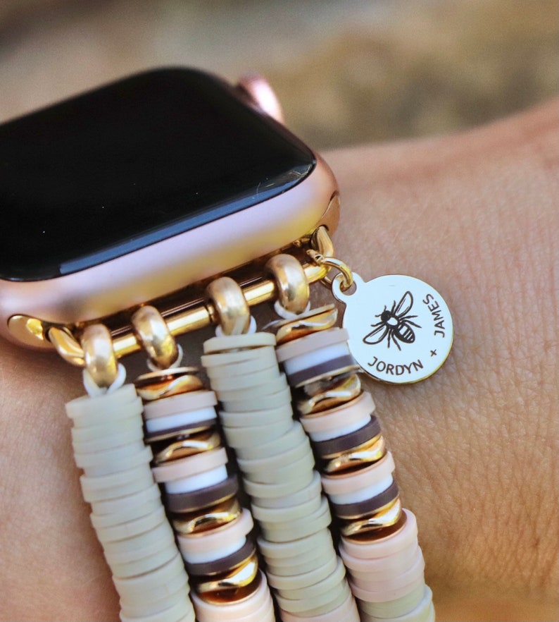 May include: A gold Apple Watch with a beige and brown beaded band. The band has a charm with a bee and the text "JAMES x JORDYN".
