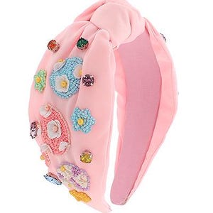 Puede incluir: Diadema de tela rosa con un lazo anudado y adornada con flores de cuentas de colores y pedrería.