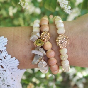 Può includere: Tre braccialetti impilati su un polso. Il braccialetto superiore è bianco con un ciondolo a forma di sfera dorata scintillante. Il braccialetto intermedio è rosa chiaro con un ciondolo a forma di sfera dorata scintillante. Il braccialetto inferiore è in legno marrone chiaro con un ciondolo dorato che dice "Magnolia Jane's".