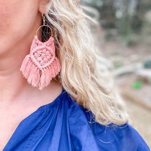 May include: Pink macrame earrings with a fringe detail. The earrings are hanging from a person's ear.