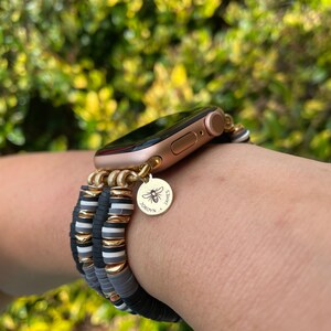May include: A gold Apple Watch with a black and white beaded bracelet. The bracelet has a gold charm with a bee and the names "Jordyn" and "James".