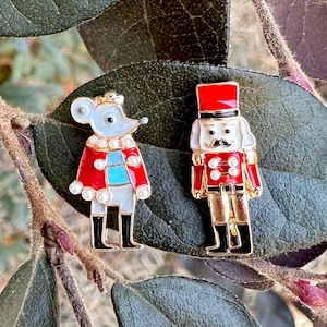 May include: A pair of gold-tone Christmas-themed enamel pins. One pin features a gray mouse wearing a red and white coat with a blue scarf. The other pin features a red and white nutcracker with a gold hat and black boots.