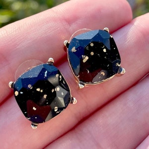 May include: A pair of gold-tone stud earrings with black square-shaped stones. The stones have a speckled gold design.