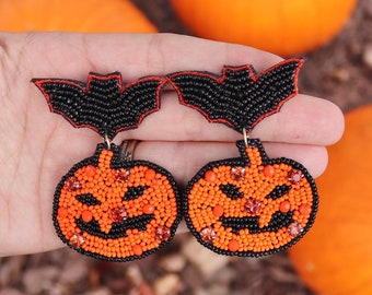 Könnte beinhalten: Ein Paar Halloween-Ohrringe mit schwarzen Fledermaus-Anhängern und orangefarbenen Kürbisperlen. Die Kürbisse haben ein Perlen-Gesicht mit schwarzen Augen und einem Mund.