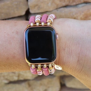 May include: A rose gold Apple Watch with a pink and white beaded band. The band is made of multiple strands of beads and has a gold charm.