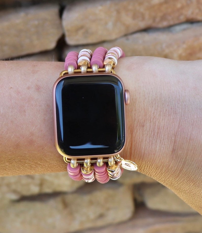 May include: A rose gold Apple Watch with a pink and white beaded band. The band is made of multiple strands of beads and has a gold charm.