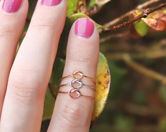 Könnte beinhalten: Drei zarte Knotenringe in Gold, Silber und Roségold werden auf einem Finger mit pinkfarbenem Nagellack präsentiert. Die Ringe sind schlicht und elegant und zeigen ein minimalistisches Design.