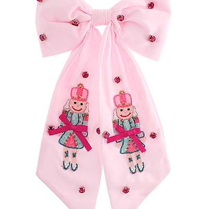 May include: A large pink bow with long streamers, decorated with embroidered nutcracker figures and sparkling red embellishments. The bow is made of a soft fabric, ideal for festive occasions.