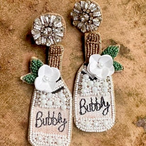 May include: A pair of champagne bottle-shaped earrings with white pearl and gold beads, a white flower, and green leaves. The earrings are decorated with the word "Bubbly" in black thread.