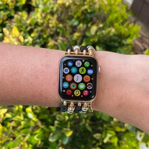 May include: A gold Apple Watch with a black and white beaded band. The watch face displays the time, date, and several app icons.