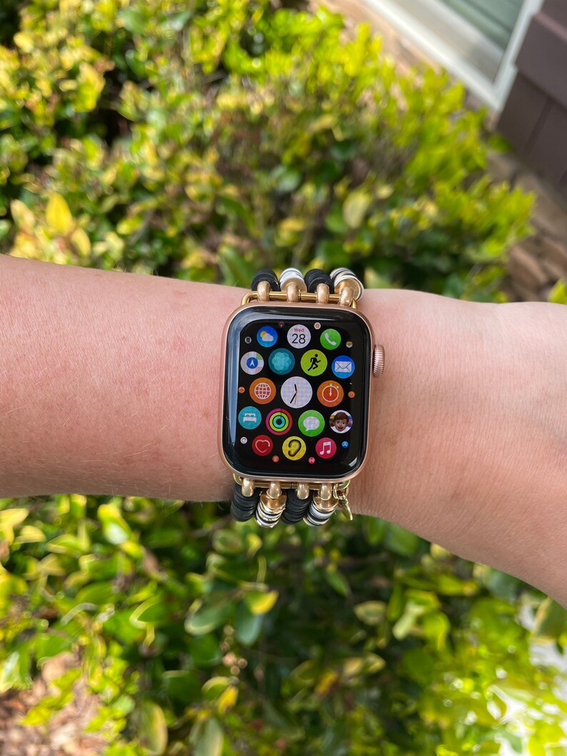 May include: A gold Apple Watch with a black and white beaded band. The watch face displays the time, date, and several app icons.