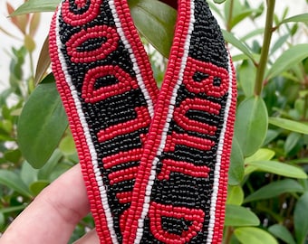 Peut inclure: Une sangle en perles rouge et noire avec les mots "GO DAWGS" et "BRING IT ON" en perles blanches.