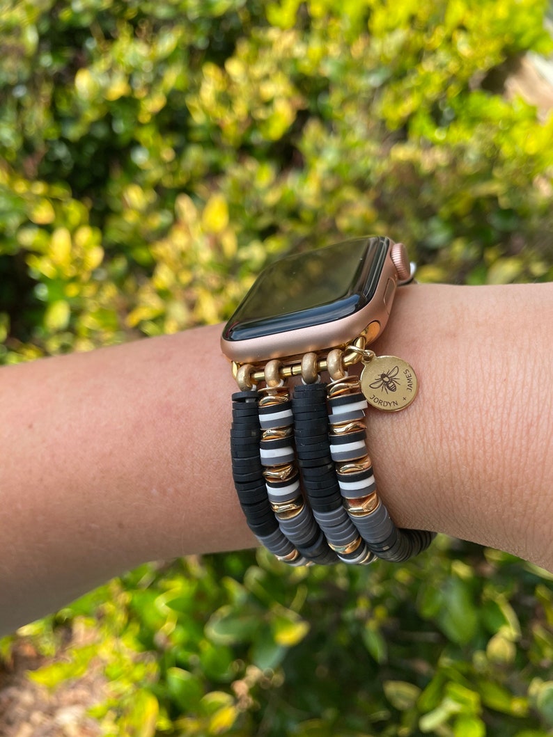May include: A gold Apple Watch with a black face and a black, white, and gray beaded bracelet. The bracelet has a gold charm with a bee and the text "JORDYN'S SWEETS".