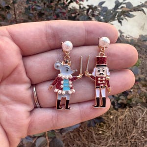 Puede incluir: Un par de pendientes festivos con un diseño de ratón y cascanueces. El ratón es gris con una corona dorada y una chaqueta roja. El cascanueces es rojo y blanco con un sombrero negro. Ambos sostienen espadas y están adornados con perlas de imitación.