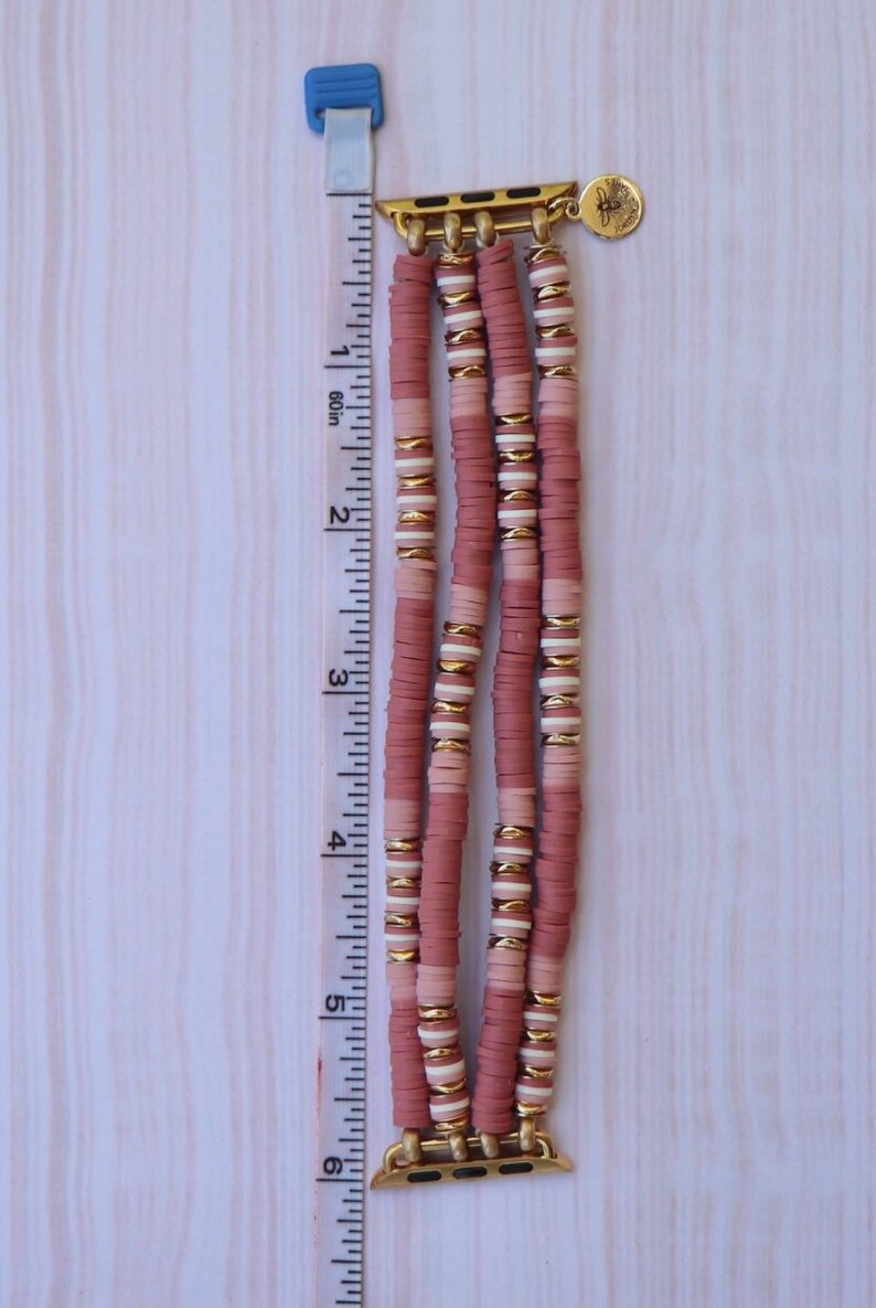 May include: A pink and white beaded Apple Watch band with gold accents. The band is made of three rows of beads and has a gold clasp.