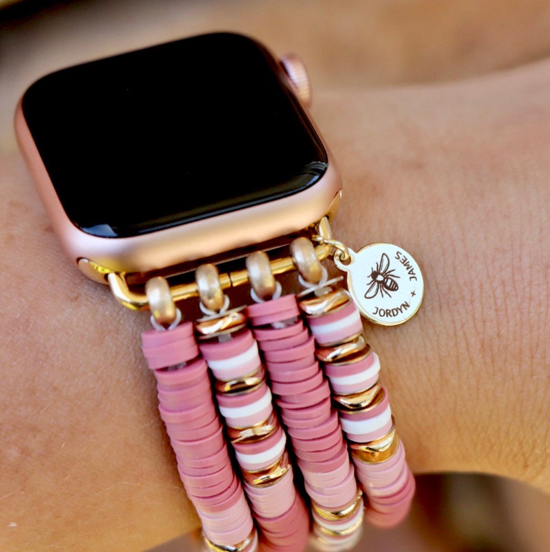 May include: A rose gold Apple Watch with a pink and white beaded band. The band has a gold charm with the text "JORDYN + JAMES" and a bee design.