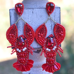 Puede incluir: Pendientes de langosta rojos de cuentas con un gran strass rojo en el centro de cada langosta. Los pendientes cuelgan de una cadena dorada.