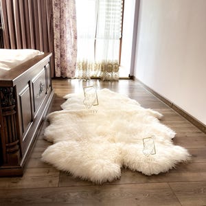 May include: A cream-colored sheepskin rug with a soft, fluffy texture lies on a hardwood floor. The rug is irregularly shaped and appears to be made of natural wool. The room has a bed and curtains in the background.