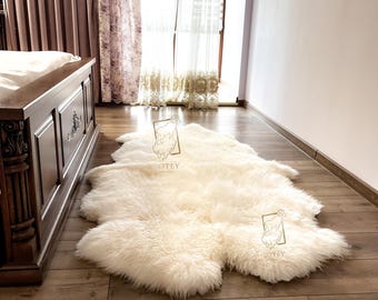 May include: A cream-colored sheepskin rug with a soft, fluffy texture lies on a hardwood floor. The rug is irregularly shaped and appears to be made of natural wool. The room has a bed and curtains in the background.