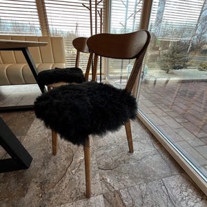 May include: Two wooden chairs with curved backs and light brown legs. Each chair has a black, fluffy seat cushion. The chairs are in front of a large window with blinds, and a table is partially visible on the left.