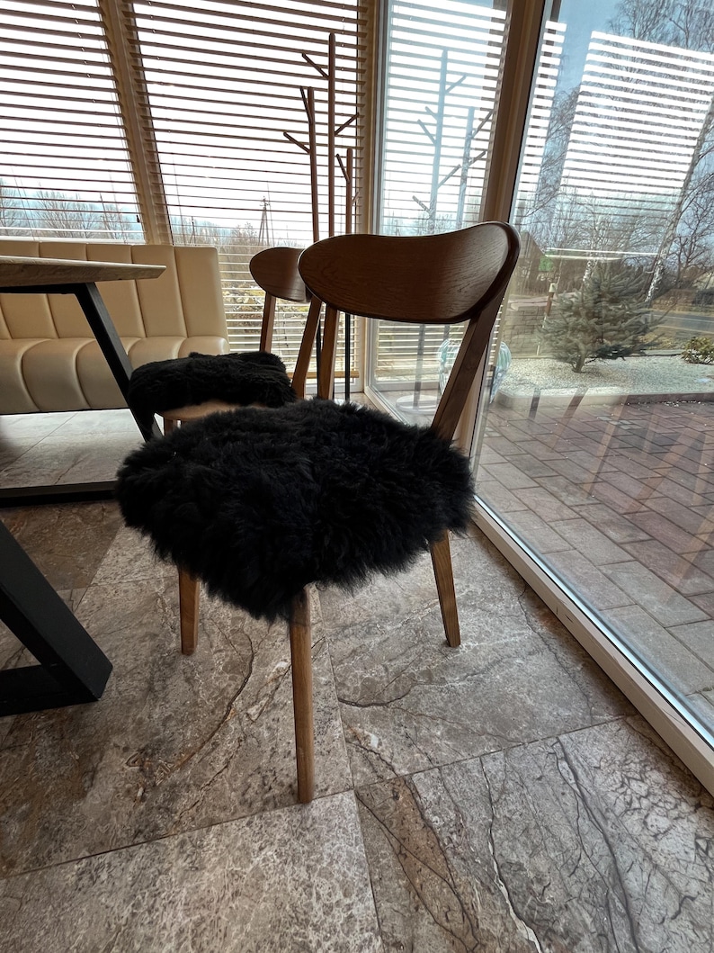 May include: Two wooden chairs with curved backs and light brown legs. Each chair has a black, fluffy seat cushion. The chairs are in front of a large window with blinds, and a table is partially visible on the left.