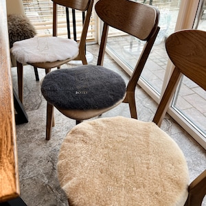 Op de afbeelding: Houten stoelen met ovale rugleuningen en ronde zittingen. Elke stoel heeft een schapenvacht zitting in wit, grijs en beige. De stoelen staan bij een raam, waardoor natuurlijk licht de ruimte verlicht.