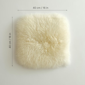 May include: Cream-colored sheepskin cushion, measuring 16 inches by 16 inches (40 cm x 40 cm). The cushion has a soft, fluffy texture and a square shape. The sheepskin is a light, natural color.