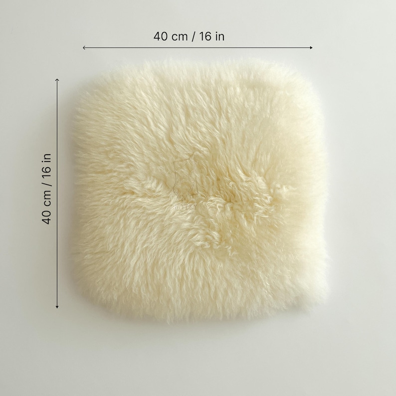 May include: Cream-colored sheepskin cushion, measuring 16 inches by 16 inches (40 cm x 40 cm). The cushion has a soft, fluffy texture and a square shape. The sheepskin is a light, natural color.