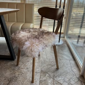 May include: A wooden chair with a curved back and four slender legs. The seat is covered with a fluffy, off-white and gray sheepskin. The chair is positioned in front of a window with blinds.
