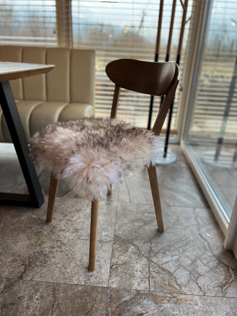 May include: A wooden chair with a curved back and four slender legs. The seat is covered with a fluffy, off-white and gray sheepskin. The chair is positioned in front of a window with blinds.