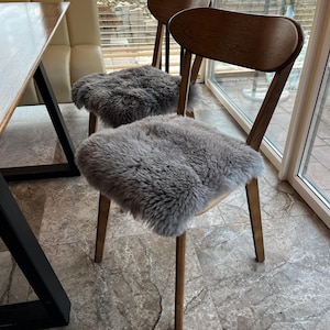 May include: Two wooden chairs with gray sheepskin seat covers. The chairs have curved backs and slender legs. The sheepskin covers add a touch of texture and comfort to the seating. The chairs are placed near a window.